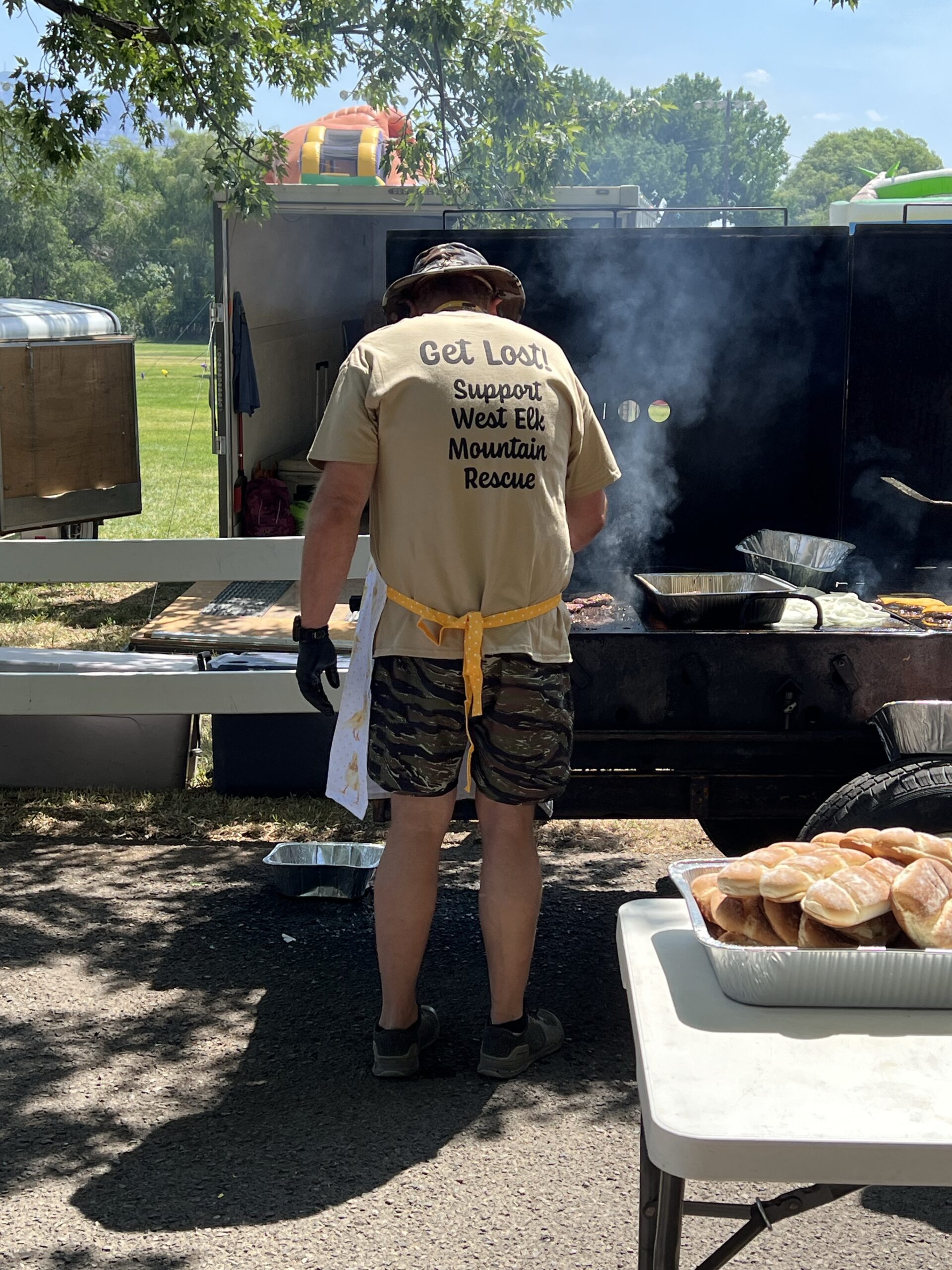A man in a Support West Elk Mountain Rescue shirt prepares burgers on a grill.